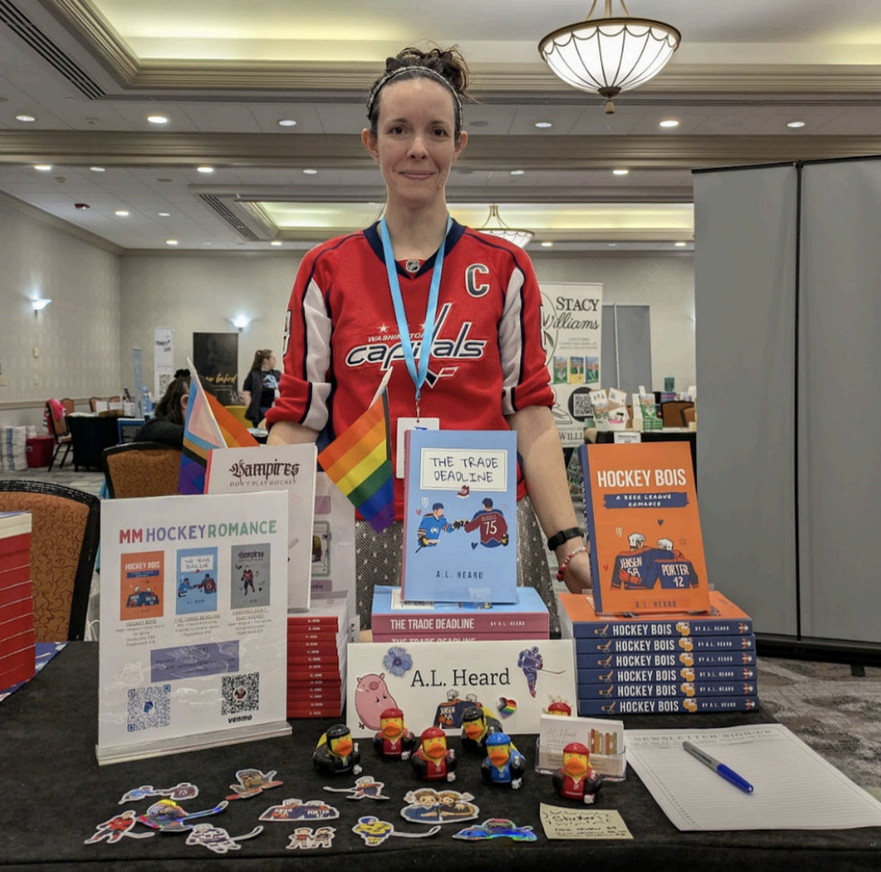 The author wears a hockey jersey and stands in front of a table. The table holds displays, stickers, and piles of books.